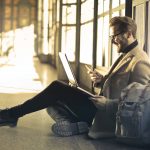 man sitting near window holding phone and laptop