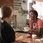 Smiling African American waitress wearing casual t shirt and apron standing at counter in cozy coffee shop with female customer while serving hot drink and looking at each other
