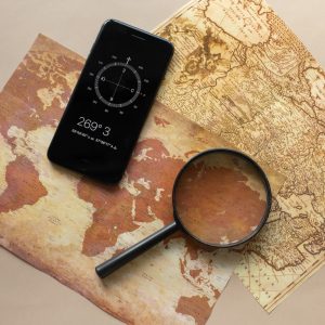Top view of magnifying glass and cellphone with compass with coordinates placed on paper maps on beige background in light room
